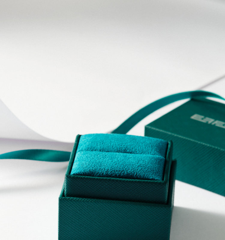 A teal velvet jewelry box with a matching ribbon sits on a white surface, partially open to reveal its cushioned interior. Light and shadows create a soft, elegant atmosphere.