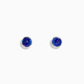 The Nahla Siri Sterling Silver Tanzanite Stud Earrings feature round deep blue tanzanite gemstones in simple sterling silver settings, shown on a white background.