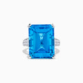 The 14K White Gold Blue Topaz and Diamond Ring, 29.29 TCW, features a large rectangular emerald-cut blue topaz accented by small white diamonds, shown on a white background.