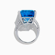 The 14K White Gold Blue Topaz and Diamond Ring, 29.29 TCW, features a large rectangular emerald-cut blue topaz accented by small white diamonds, shown on a white background.