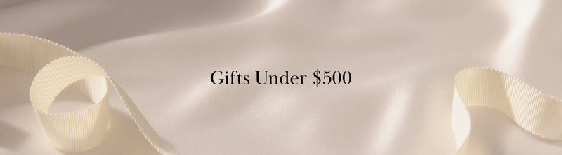 A cream-colored ribbon lies on smooth, light fabric with the text Gifts Under $500 centered in elegant black font.
