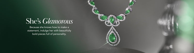 Close-up of an elegant necklace with green gemstones and diamonds on a display, beside text: Shes Glamorous. Because she knows how to make a statement, indulge her with beautifully bold pieces full of personality.