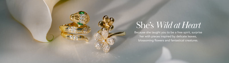 Three gold rings with floral and leaf designs, some adorned with green and clear stones, are displayed on a soft surface next to a white flower, with the text: She’s Wild at Heart.