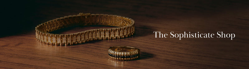 A gold bracelet and ring with intricate designs rest on a polished wooden surface next to the text “The Sophisticate Shop.”.