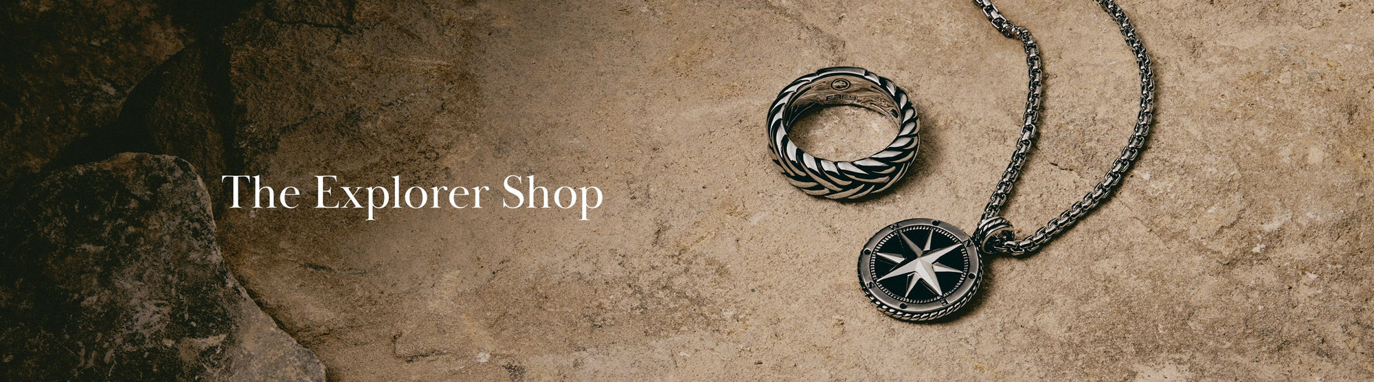 A silver ring and a compass pendant necklace rest on a textured stone surface. Text reads The Explorer Shop on the left side.