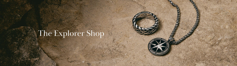 A silver ring and a compass pendant necklace rest on a textured stone surface. Text reads The Explorer Shop on the left side.