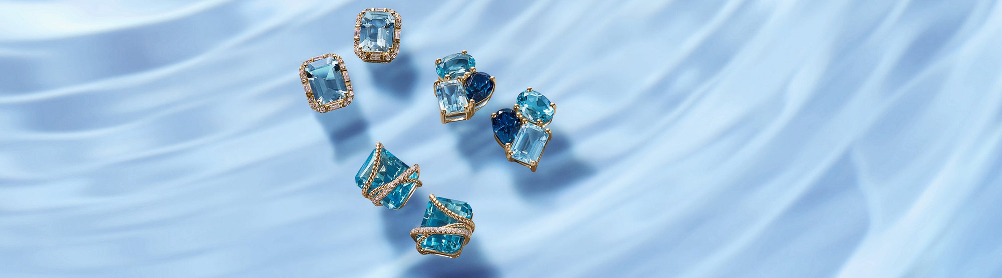 Six gold-edged blue topaz and aquamarine gemstone rings on a silky, light blue fabric background with the text “Blue Topaz & Aquamarine” on the right.
