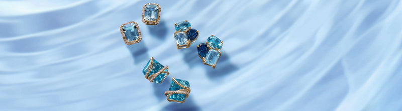 Six gold-edged blue topaz and aquamarine gemstone rings on a silky, light blue fabric background with the text “Blue Topaz & Aquamarine” on the right.