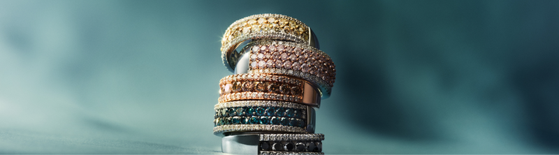 A stack of sparkling gemstone and diamond rings in gold, rose gold, and silver finishes on a blue background, with the text Best Sellers on the left.
