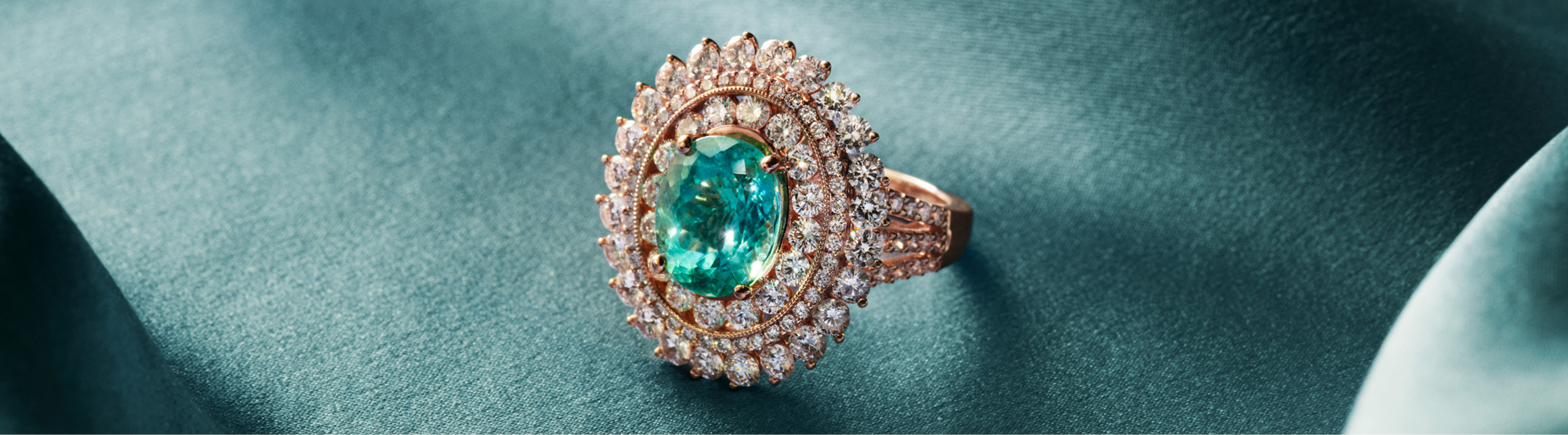 A gold ring with a large green gemstone surrounded by diamonds rests on a silky teal fabric. The text Exotic Gems appears on the left side.