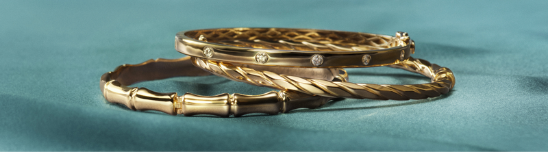 Three elegant gold bangles, one set with diamonds, rest on a soft teal fabric surface. The word Gold appears to the left in white text.