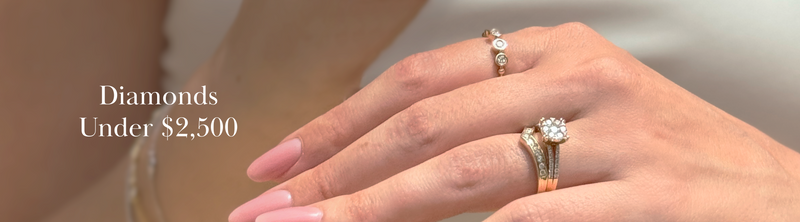 A close-up of a womans hand wearing three diamond rings, with the text Diamonds Under $2,500 displayed on the left side against a soft background.