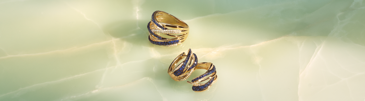 Three gold rings with intertwined bands featuring blue and white stones are displayed on a smooth, light green marble surface.