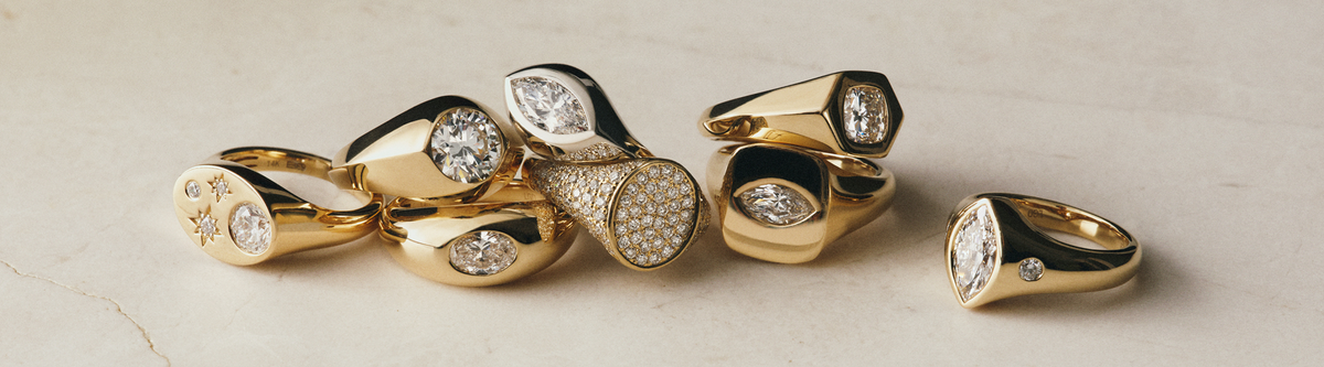 A group of gold signet rings with various diamond and gemstone settings displayed on a light marble surface.