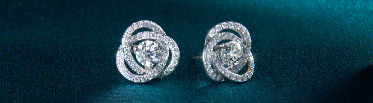 Close-up of a pair of diamond stud earrings with intricate swirling designs, set in white metal, displayed on a teal textured surface.