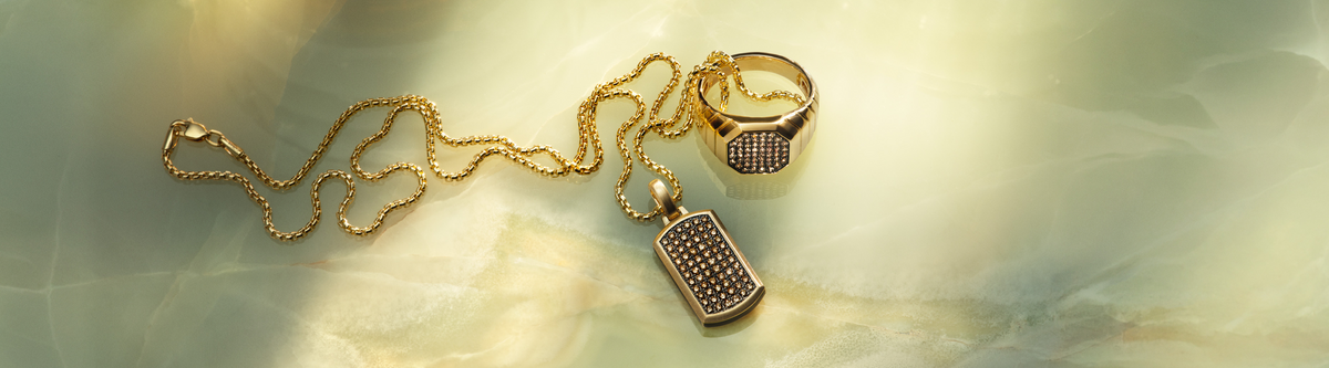 Gold chain necklace and matching gold ring with a textured, patterned design rest on a light green marble surface, illuminated by soft, natural light.