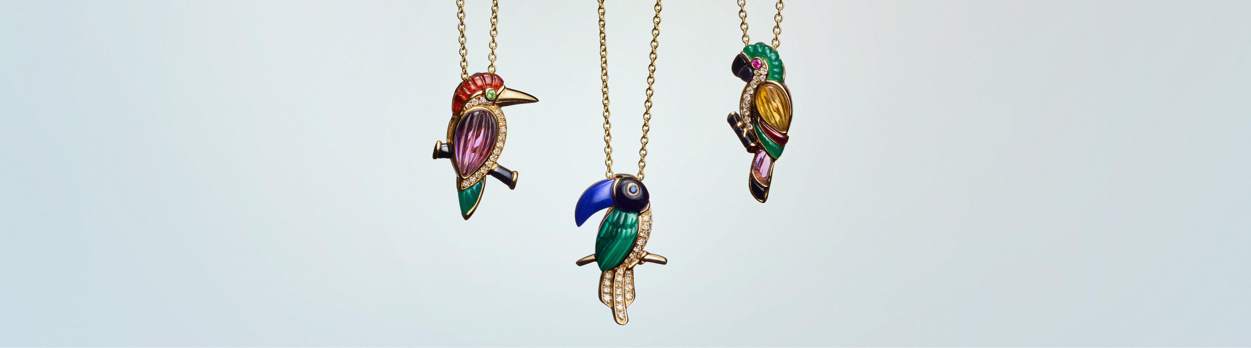 Three jeweled bird pendants on gold chains are displayed against a light background, each featuring vibrant colored stones and intricate detailing.