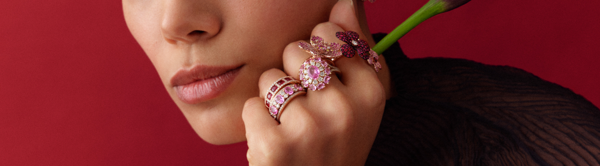 A woman with natural makeup poses against a red background, wearing bold pink gemstone rings shaped like flowers and butterflies. She holds a purple calla lily near her chin.