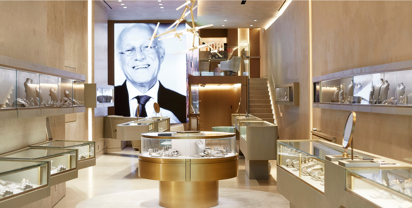 A modern jewelry store interior with glass display cases, elegant lighting, and a large black-and-white portrait of a smiling man on the back wall. A staircase is visible at the back of the store.