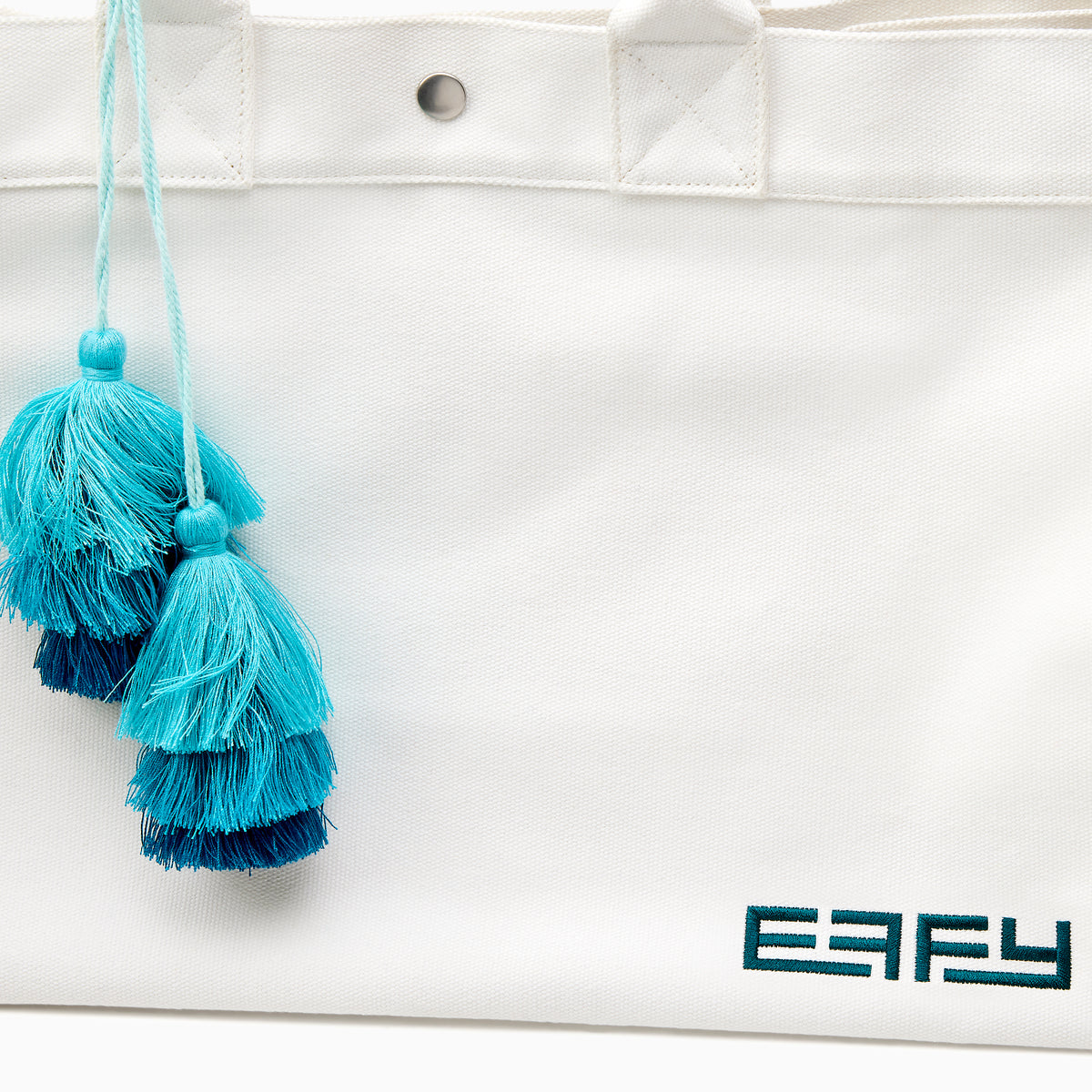 The Beach Bag - White is a large canvas tote with two handles, featuring blue and turquoise tassels and EAFY printed in black near the bottom right corner.
