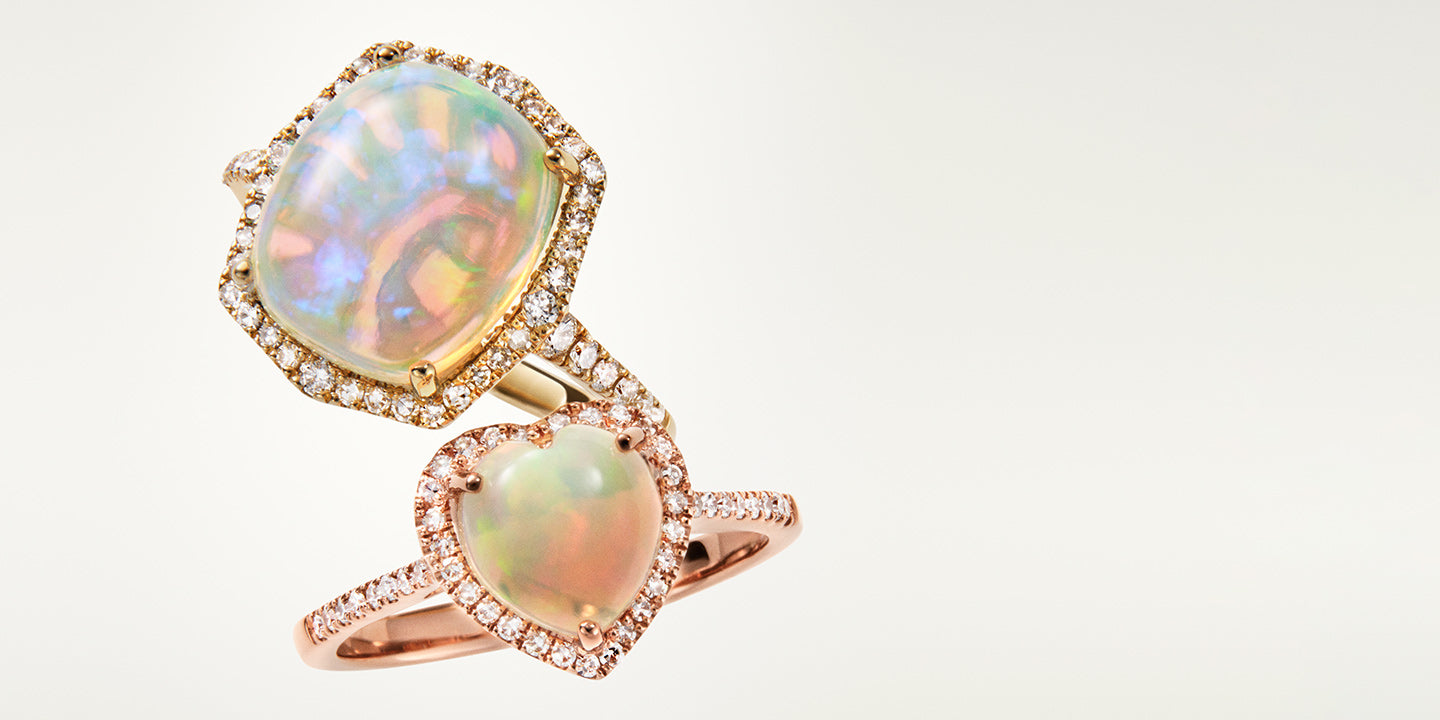 Two rose gold rings, one with a large oval opal surrounded by small diamonds, the other with a heart-shaped opal also bordered by diamonds, displayed against a soft, light background.