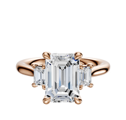 The 14K Rose Gold 3-Stone Emerald Cut Side Stones 0.125TCW Engagement Ring features a central emerald-cut diamond with two smaller rectangular diamonds on each side, displayed against a plain white background.