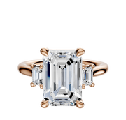 The 14K Rose Gold 3-Stone Emerald Cut Side Stones 0.25TCW Engagement Ring features a large emerald-cut center stone with two rectangular clear side stones, all set in a classic prong setting.
