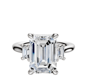 An 18K white gold engagement ring with a three-stone design, featuring an emerald-cut center stone and two rectangular side stones totaling 0.25TCW, set on a polished band.