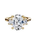The 18K Yellow Gold 3-Stone Oval Side Stones 0.125TCW Engagement Ring features a central oval diamond flanked by smaller round diamonds in a classic prong setting, displayed against a white background.