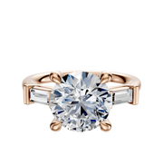 The 14K Rose Gold 3-Stone Baguette East/West Side Stones 0.125TCW Engagement Ring features a large round diamond in a four-prong setting, accented by two baguette-cut side stones, shown on a plain white background.