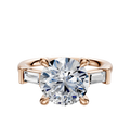 An 18K rose gold engagement ring featuring a round center stone and east/west baguette side stones (0.125TCW), shown on a plain white background.