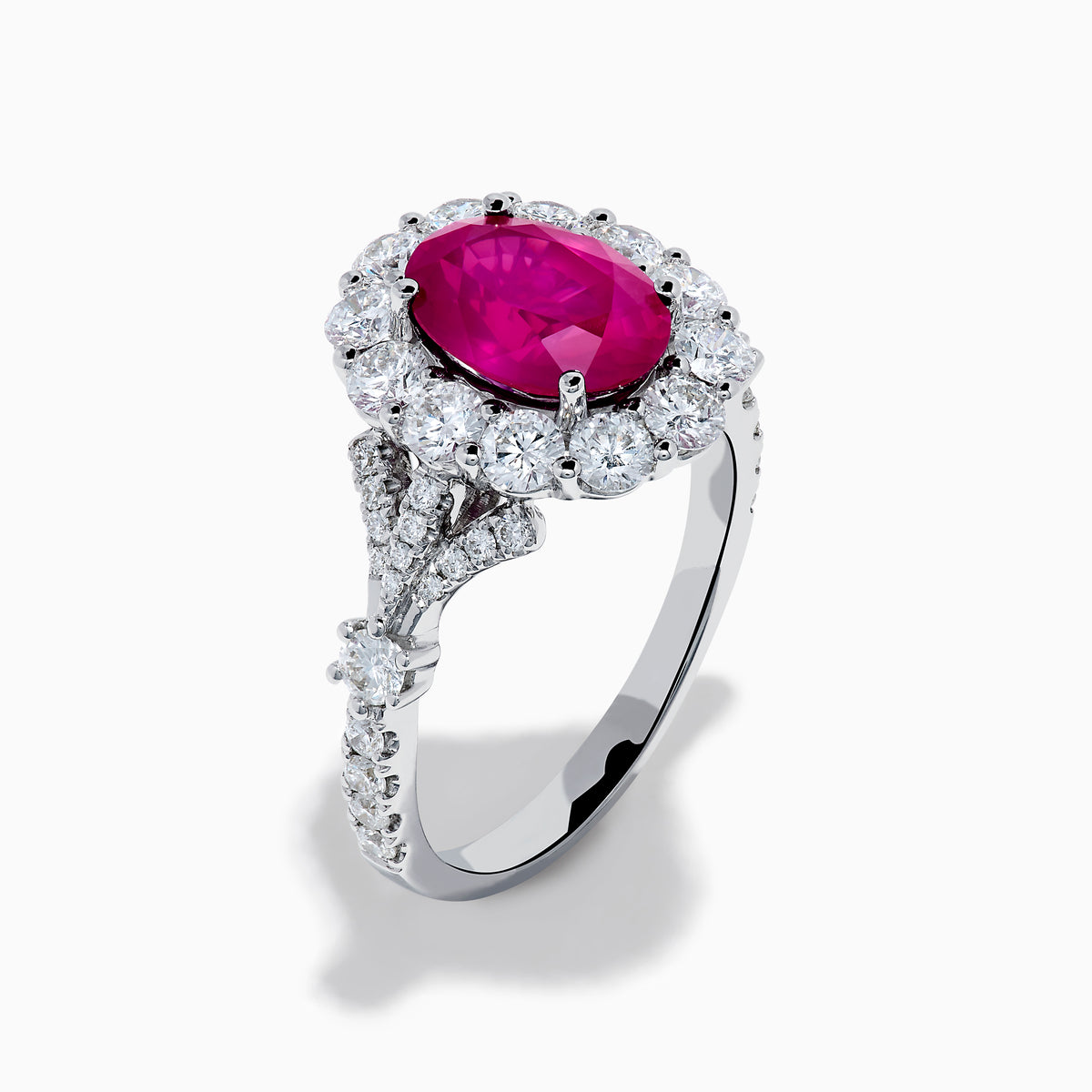 The 18K White Gold Ruby and Diamond Halo Ring features an oval red ruby at the center, encircled by a diamond halo, beautifully presented on a plain white background.