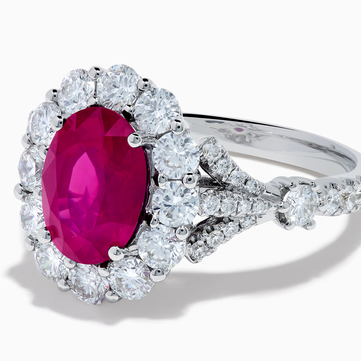 The 18K White Gold Ruby and Diamond Halo Ring features an oval red ruby at the center, encircled by a diamond halo, beautifully presented on a plain white background.