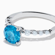 The Lab Grown Blue and White Diamond Ring set in 14K White Gold features a large round blue and white diamond at the center, accented by smaller clear stones, displayed against a white background.