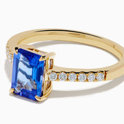 The Nahla 14K Yellow Gold Tanzanite and Diamond Ring showcases a rectangular blue tanzanite center stone, accented by small round white diamonds along the upper band on a plain white background—timeless elegance in every detail.