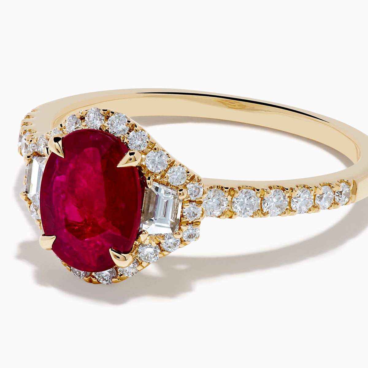 The Ruby Royale 14K Yellow Gold Ruby and Diamond 3 Stone Ring showcases a central oval ruby flanked by sparkling diamonds, set on a diamond-accented band and elegantly displayed against a white background.