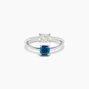 The 14K White Gold Blue and White Diamond Split Shank Ring features a clear oval and blue cushion-cut gemstone set side by side on a minimalist band, showcasing striking contrast between the blue and white diamond accents.