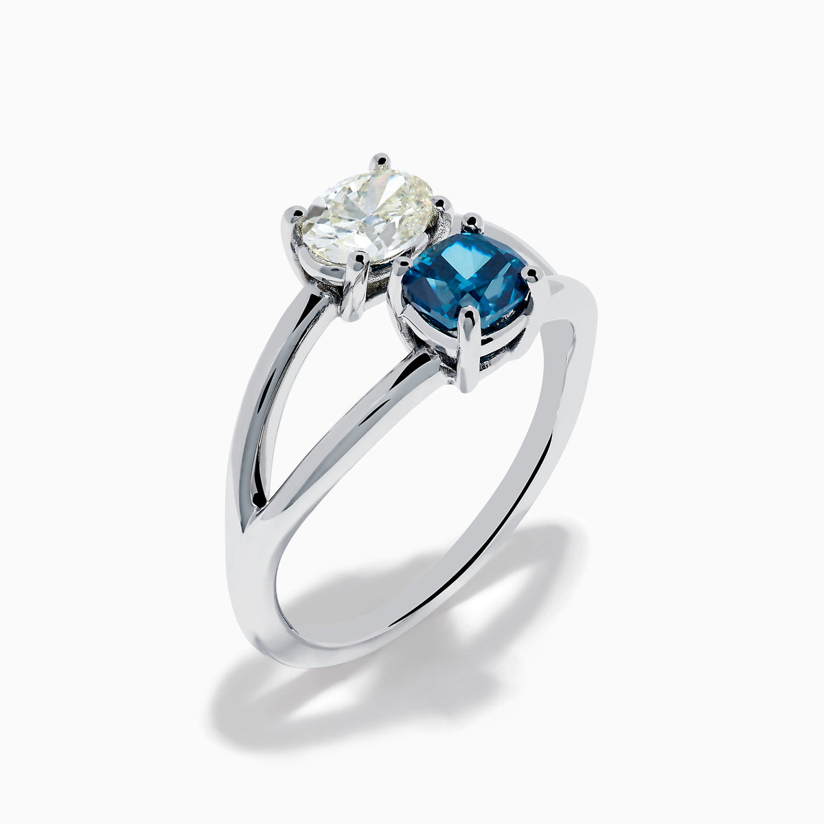 The 14K White Gold Blue and White Diamond Split Shank Ring features a clear oval and blue cushion-cut gemstone set side by side on a minimalist band, showcasing striking contrast between the blue and white diamond accents.