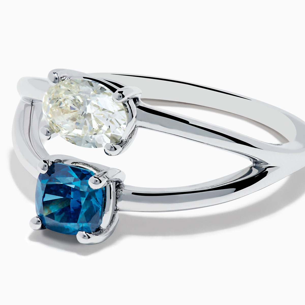 The 14K White Gold Blue and White Diamond Split Shank Ring features a clear oval and blue cushion-cut gemstone set side by side on a minimalist band, showcasing striking contrast between the blue and white diamond accents.
