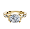 A 14K yellow gold engagement ring featuring a flat cathedral band with a 2.5mm oval center stone in an east/west half bezel and one row of pave diamonds along the band.