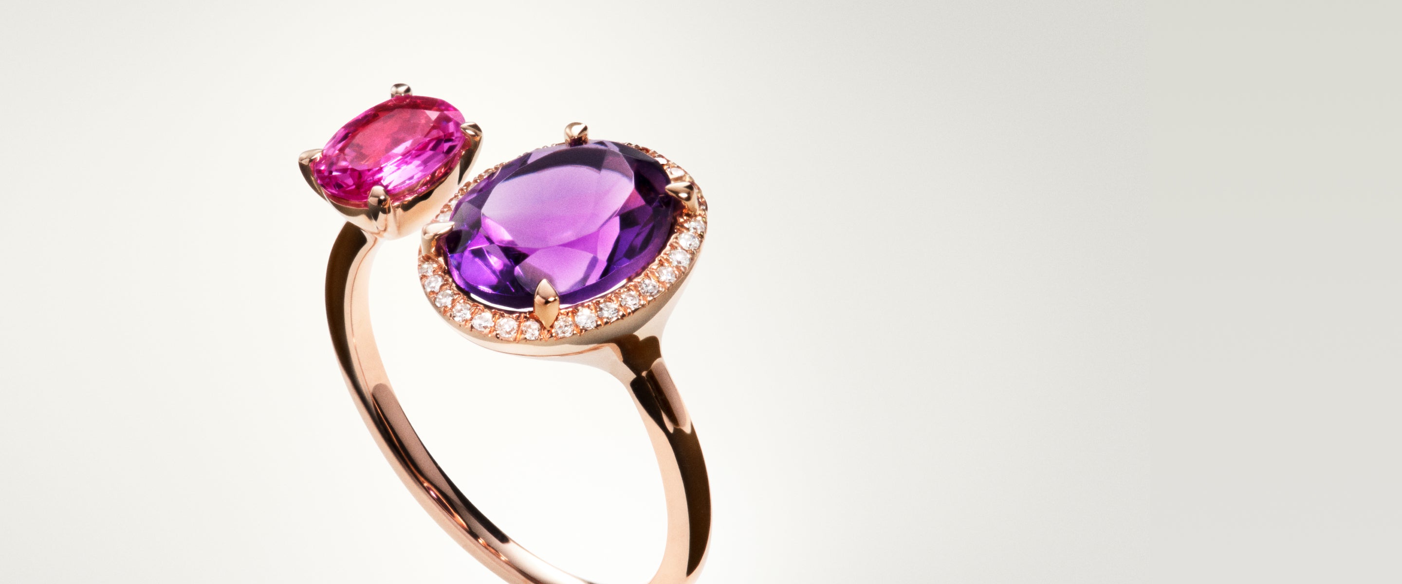 A rose gold ring featuring two gemstones: a small oval pink stone and a larger round purple stone surrounded by small diamonds, set against a plain light background.
