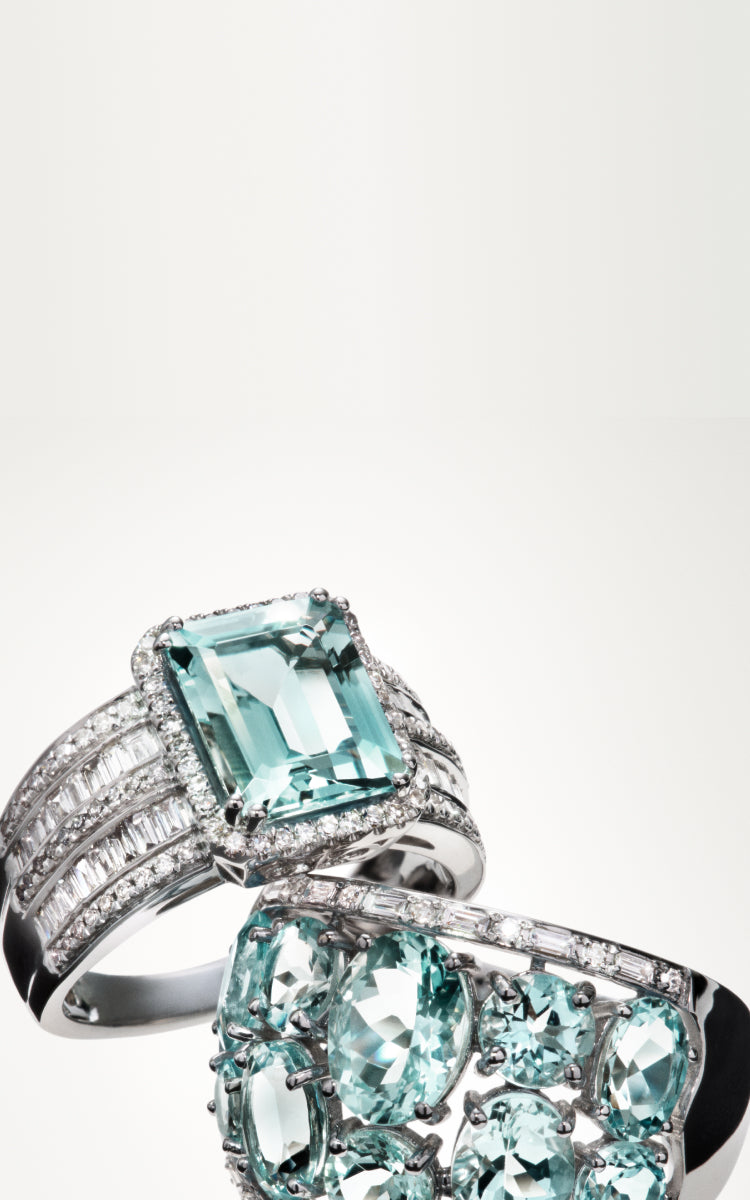 Two elegant silver rings adorned with large, pale blue gemstones and sparkling diamonds are displayed against a plain, light background. One ring features a rectangular gemstone, while the other showcases multiple oval stones.
