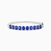 The 14K White Gold Tanzanite and Diamond Bangle features a row of oval-shaped blue tanzanites set in the center, displayed against a white background.