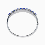 The 14K White Gold Tanzanite and Diamond Bangle features a row of oval-shaped blue tanzanites set in the center, displayed against a white background.