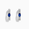 The 14K White Gold Blue Sapphire and Diamond Hoops feature an oval blue sapphire at the center, accented by rows of sparkling diamonds. Displayed on a plain white background.