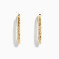 The 14K Yellow Gold Crystal Cut Tube Oval Hoops feature a textured, engraved floral pattern and delicate crystal cut detailing, shown against a plain white background.
