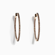 The 14K Rose Gold Brown Diamond 1.5 Inch Hoop Earrings, featuring slim vertical hoops with dark brown diamonds, are photographed against a plain white background, casting soft oval-shaped shadows to the left.