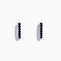 The Royale Bleu 14 Karat White Gold Blue Sapphire Huggie Hoop Earrings feature a row of round, dark blue sapphires and are shown against a plain white background.