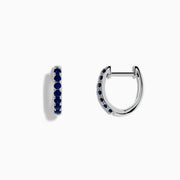 The Royale Bleu 14 Karat White Gold Blue Sapphire Huggie Hoop Earrings feature a row of round, dark blue sapphires and are shown against a plain white background.