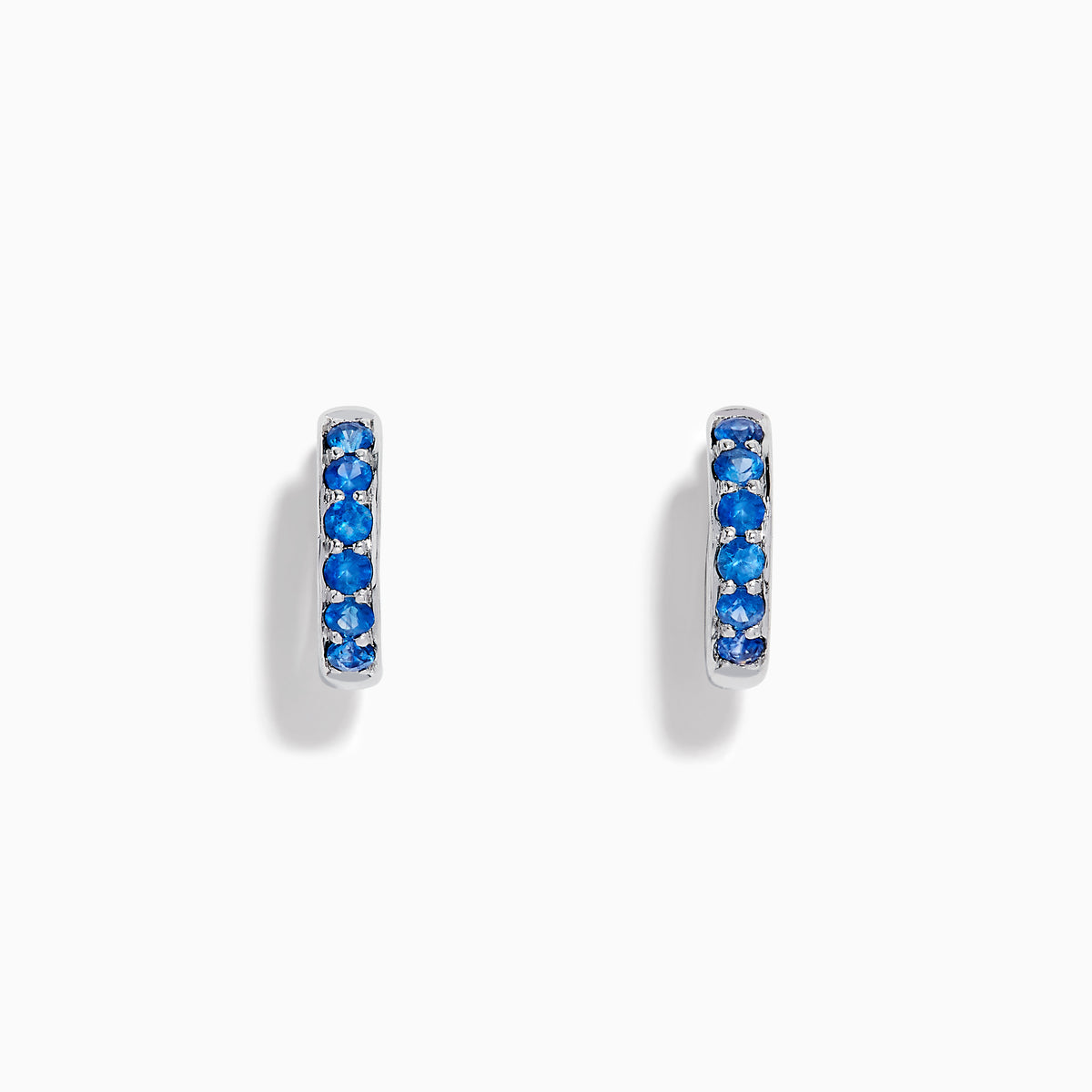 The Royale Bleu 14K White Gold Sapphire Huggie Earrings feature a row of round blue sapphires and are displayed against a white background.